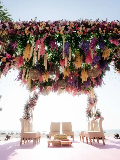 A breathtaking beachside mandap in Thailand, adorned with a canopy of cascading purple, pink, and yellow flowers, perfectly framing the ocean horizon.