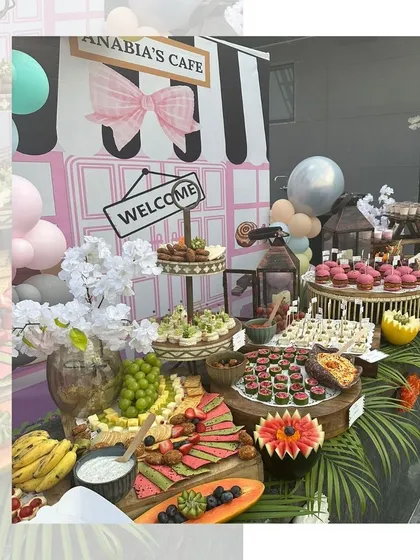 A beautiful, café themed grazing table for a little one's party. The setup includes pink cupcakes, fruit platters, and other sweet treats arranged against a charming illustrated backdrop.