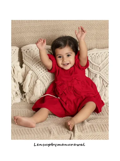 A joyful toddler in a vibrant red dress sits on a textured boho-style sofa, her arms raised in pure happiness, capturing the uncontainable energy of childhood.