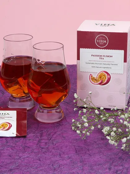 A lifestyle product photograph for Passion Fusion Tea. This setup includes the product box, individual tea sachets, and two glasses of the brewed beverage, arranged on a textured purple surface with delicate flowers.