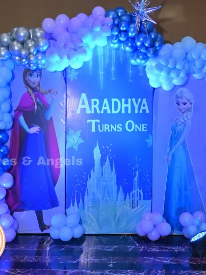 A magical Frozen theme for Aradhya's first birthday. The backdrop features Elsa and Anna against an icy castle, framed by a blue, purple, and silver balloon arch to create a winter wonderland.