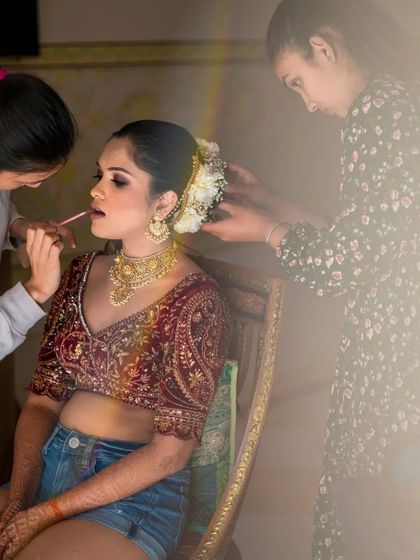 A candid shot of the bride getting her makeup done, casually dressed in jeans with her elaborate blouse and jewelry, showing a real behind the scenes moment.