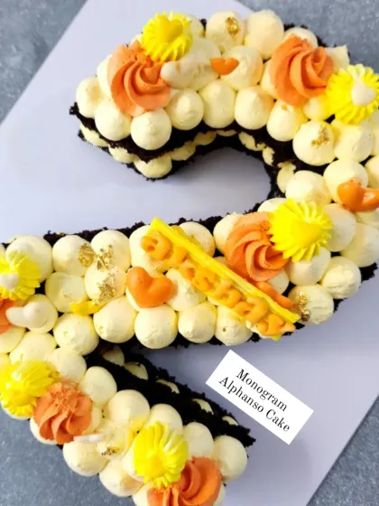 A monogram "S" cake made with a chocolate base and piped with fresh Alphonso mango cream, decorated with orange and yellow rosettes.
