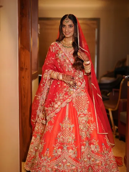 A full-length view of the bride in her stunning red-orange lehenga. The makeup is kept soft and glowing to balance the vibrant color of her outfit.