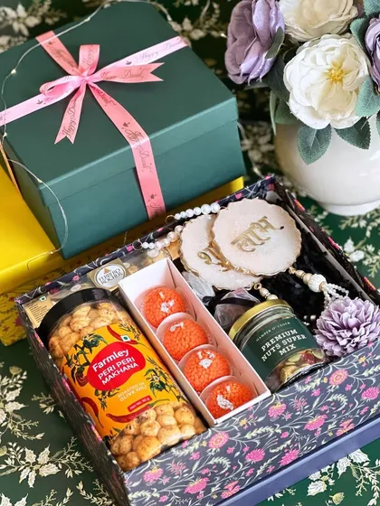 This beautifully arranged Diwali hamper features a dark floral print box. Inside, it offers a mix of savory and sweet, with peri-peri makhana, ladoo candles, a premium nut mix, and Shubh Labh hangings for good fortune.