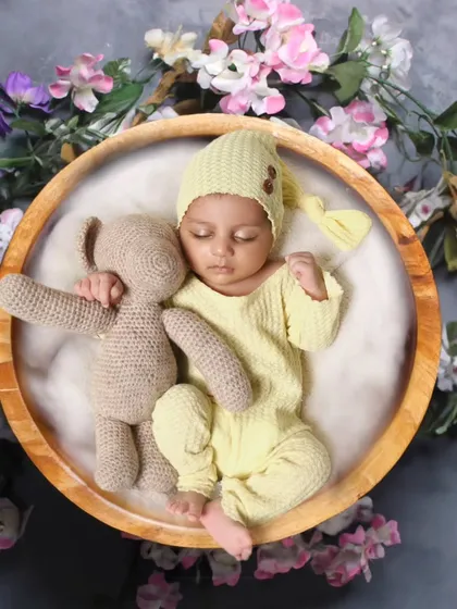 A 15-day-old baby boy in a yellow outfit, sleeping with a teddy bear in a wooden bowl surrounded by flowers.