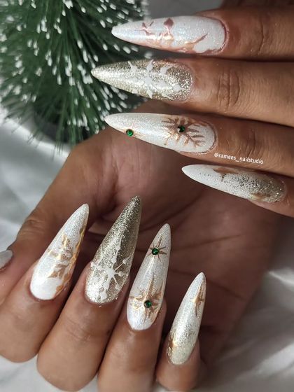 A dramatic and festive Christmas look. These long stiletto nails feature a white and gold glitter base with snowflake designs and green rhinestone accents.