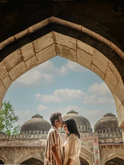Framing the couple within a stone archway, with the domes of a historic monument visible in the background. This is a classic pre-wedding shot that combines romance with architectural beauty.