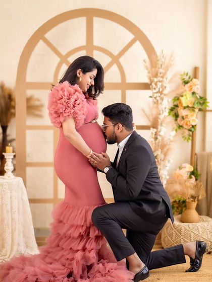 A tender moment between an expecting couple, with the dad-to-be kissing his partner's baby bump. Her ruffled pink mermaid gown adds a touch of modern romance.