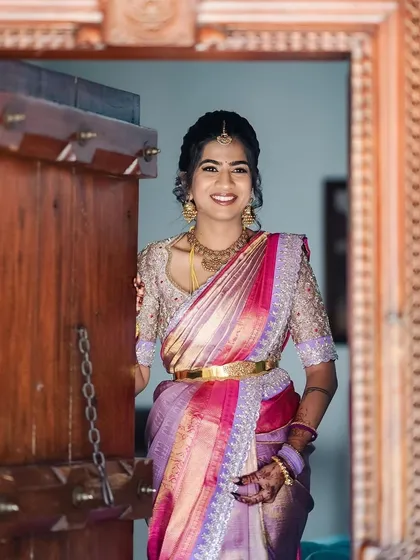 A full view of the bride in her pink and lavender saree, standing in a traditional doorway. The intricate blouse and rich saree colors make for a stunning bridal portrait.