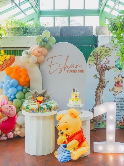 A beautiful setup for Eshan's "Bee ONE!" Winnie the Pooh party. The scene is set in a bright, plant-filled room, featuring a large Pooh Bear prop, a "Hundred Acre Wood" sign, and a marquee number '1'.