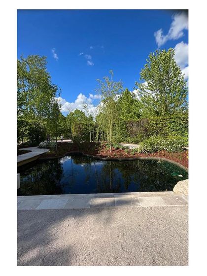 The dark, reflective surface of the pond mirrors the bright blue sky and surrounding trees. Water is a central element in our designs, bringing life, reflection, and a sense of calm to the landscape.