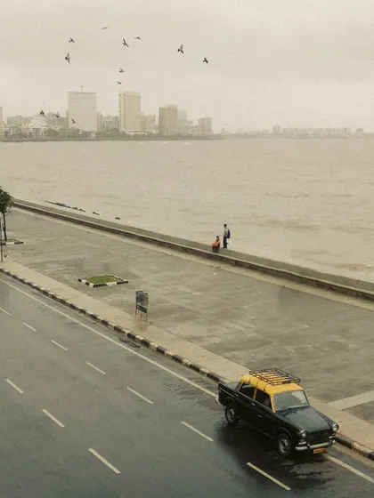 An elevated view of a lone Kaali-Peeli taxi on the road beside Marine Drive. The composition contrasts the small taxi with the vast sea and the distant skyline, creating a sense of scale and journey.