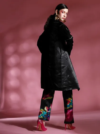 A back view of a model wearing a black coat over colorful printed trousers. The styling with bright pink heels adds an unexpected pop of color, completing the look.