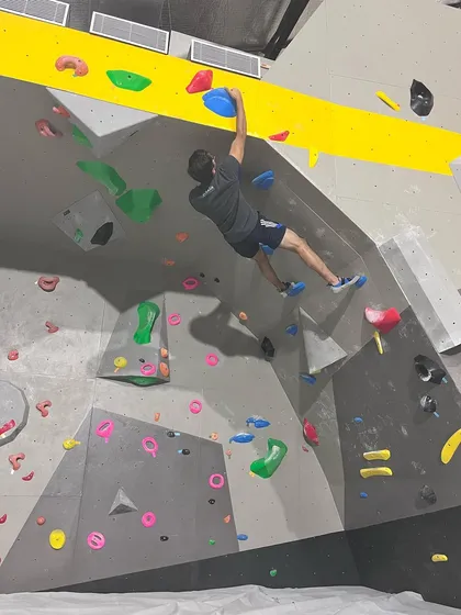 A climber reaches high on a blue hold, demonstrating the full-body extension needed for certain routes. Our walls have problems designed for all heights and reach.