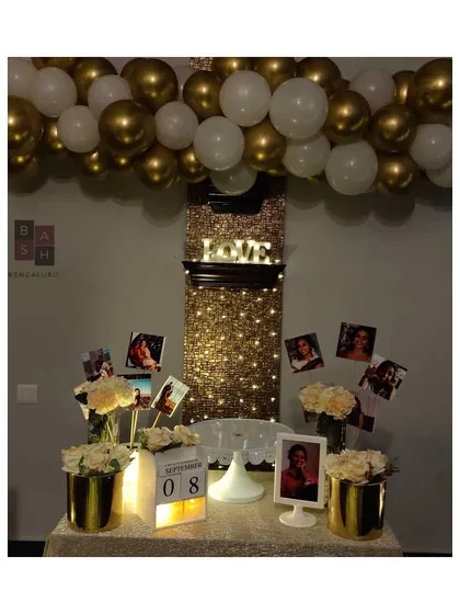 We can transform even a simple wall into a stunning feature. This date night setup includes a shimmering curtain of fairy lights, a personalized photo display table, and a beautiful gold and white balloon garland.