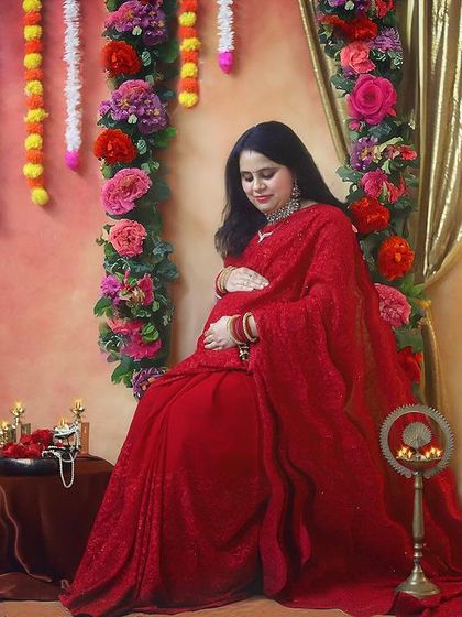 A serene moment of reflection. This seated pose in a gorgeous red saree, surrounded by flowers and traditional decor, captures the quiet joy of waiting for your little one.