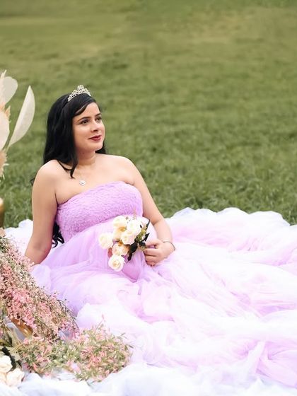 A fairytale-like outdoor portrait. The mother-to-be, wearing a tiara and a flowing lavender gown, is seated on a picnic blanket surrounded by whimsical props like white pumpkins and pampas grass.