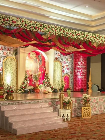 This stage is a beautiful example of traditional Indian wedding decor. It features a backdrop with classic motifs, draped in red and gold fabrics, and a wooden mandap structure adorned with florals, creating a rich, cultural ambiance.