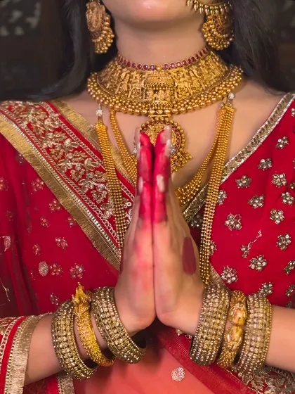 A close-up shot focusing on the traditional elements of the Padmavati look. The hands are held in a namaste gesture, adorned with alta and heavy gold bangles, with the temple necklace visible.