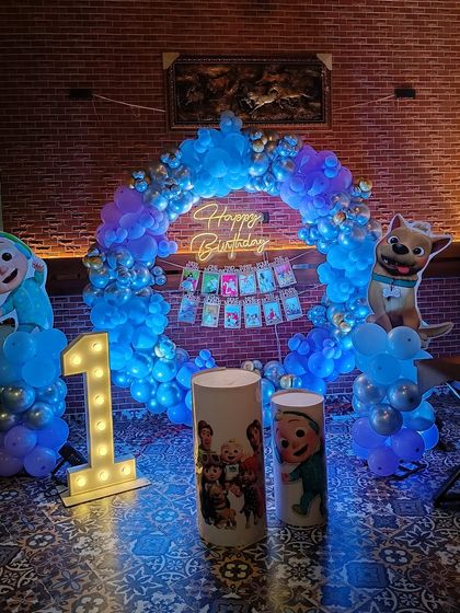 A Cocomelon-themed first birthday party perfect for toddlers. This setup includes a blue balloon ring, character cutouts, themed pedestals, and a light-up number '1'.