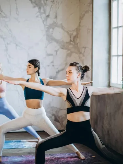 Three students demonstrate Virabhadrasana II (Warrior II). The caption details the alignment cues and benefits, such as stretching the hips and improving stamina.