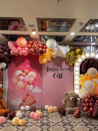 A warm and inviting teddy bear theme for a first birthday. This setup uses a rich color palette of brown, gold, and cream balloons, with multiple teddy bears and two illuminated arch backdrops.