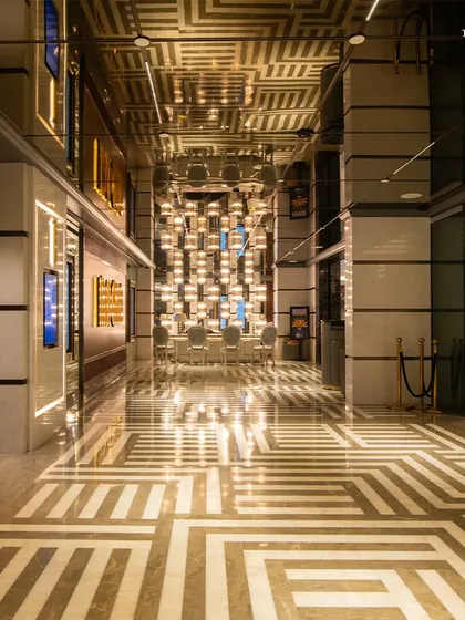 A wide shot of the grand corridor at Maison INOX, where geometric patterns on the floor and ceiling create a mesmerizing effect. The design uses reflection, light, and opulent materials to transport visitors to the golden age of cinema.