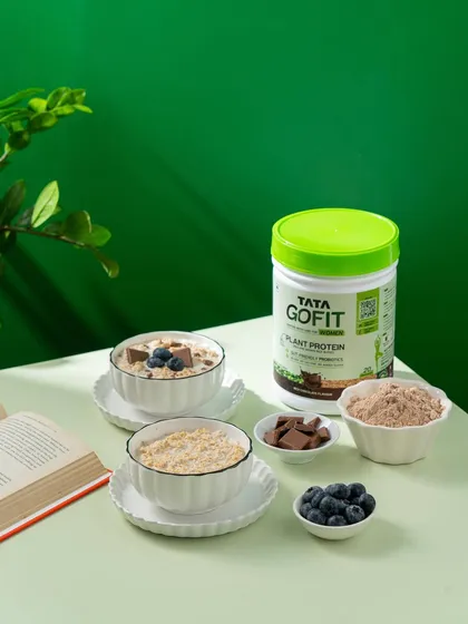 A wider angle of the healthy breakfast scene, showing the Tata GoFit protein powder alongside bowls of oats, blueberries, and chocolate.