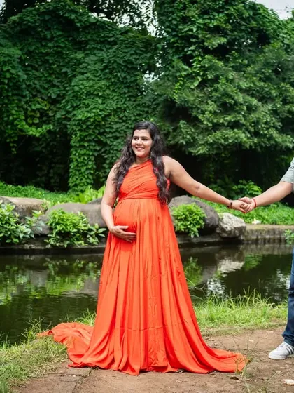 A wide shot showing the mom-to-be in a vibrant orange gown, holding hands with her partner by a pond. It's a beautiful, scenic maternity portrait.