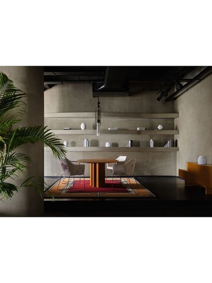 A wider view of the dining setup, showing how the colorful rug anchors the space. The design uses a mix of materials, from the metal legs of the chairs to the wooden table and concrete-finish walls.