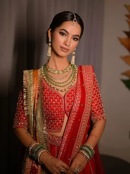 A confident and regal pose from the bride. Her makeup features strong, defined brows and contoured cheeks, balancing the grandeur of her multi-layered necklaces and traditional red attire.