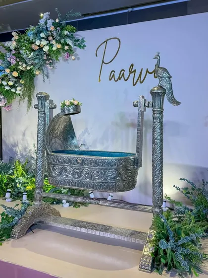 The silver cradle for Paarvi's ceremony, adorned with a peacock motif and set against a clean white backdrop with pastel floral arrangements.