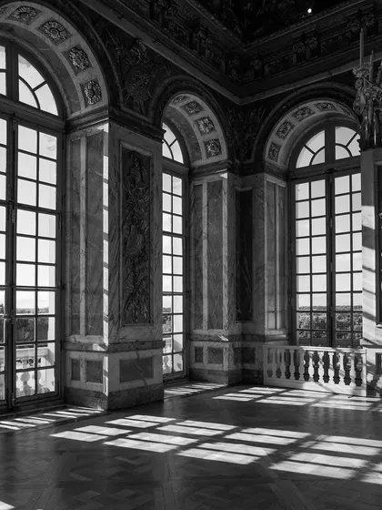 Light streams through the grand windows of the Palace of Versailles, casting geometric shadows on the floor. Black and white emphasizes the stunning play of light and architecture.