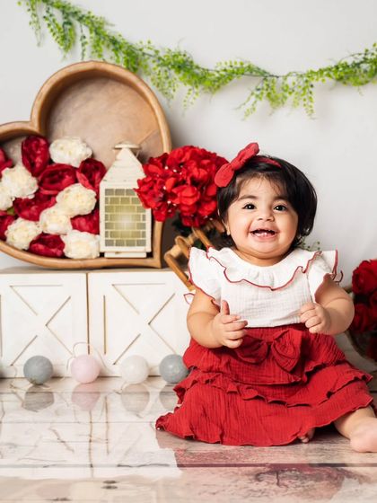 The birthday girl gives a big, joyful smile. Her red dress and bow perfectly match the romantic, floral-themed first birthday set.