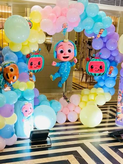 A vibrant Cocomelon-themed birthday party. This setup features a pastel-colored balloon arch, multiple character cutouts and foil balloons, and themed pedestals.