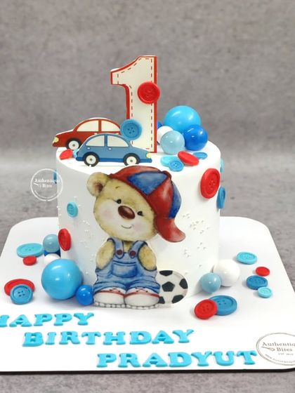 A playful first birthday cake featuring a teddy bear, cars, and colorful buttons. A perfect design for a little boy's special day.