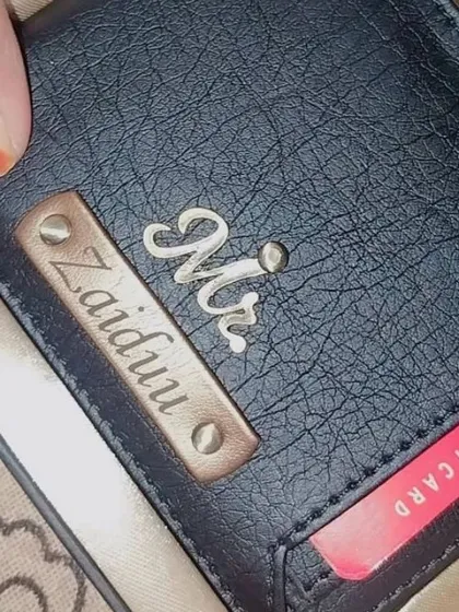 A close-up view of the black personalized wallet with the red credit card slot, showing the detail of the name engraving and charm.