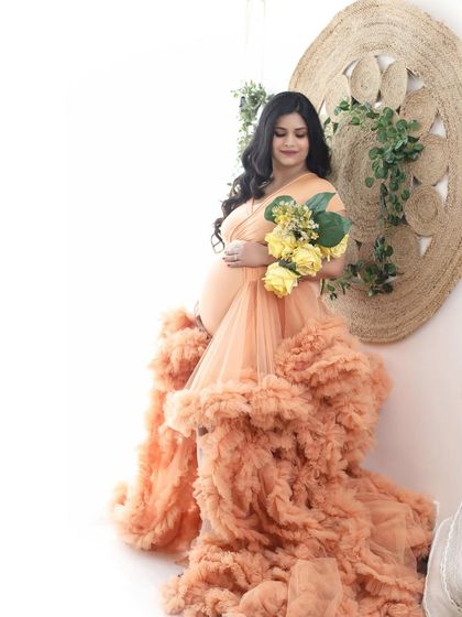 A dreamy, artistic portrait of a mom-to-be in a peach ruffled gown, holding a bouquet of yellow flowers. The bright, clean background enhances the ethereal feel.