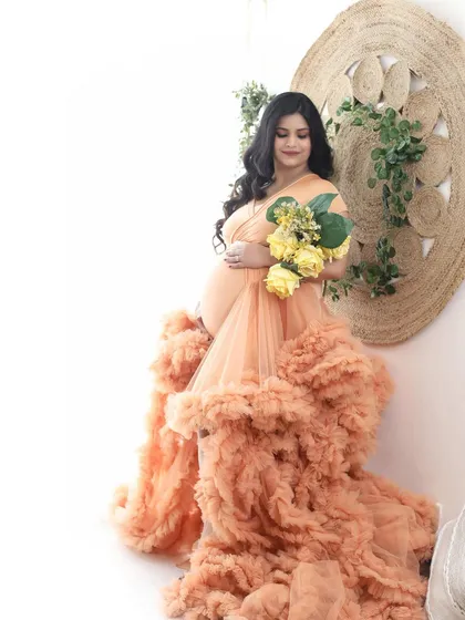 A dreamy, artistic portrait of a mom-to-be in a peach ruffled gown, holding a bouquet of yellow flowers. The bright, clean background enhances the ethereal feel.