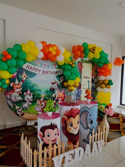 A classic jungle book style setup for a first birthday. It includes a circular backdrop with cartoon animals, character-themed pedestals, and a colorful balloon arch.