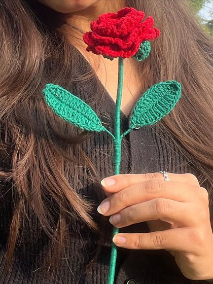 A close-up of a single, perfect red crochet rose held by a model. The detail in the petals is a testament to its handmade quality.