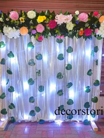 A nature-inspired ready-to-install backdrop for Ganpati. This design features cool white lights behind a sheer curtain, decorated with green leaf creepers and a colorful floral border at the top.