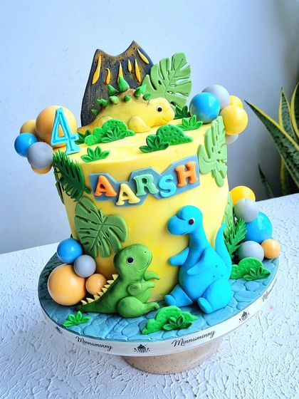 A bright yellow dinosaur cake with a volcano and several cute fondant dinosaurs. The balloon bunches are made from chocolate spheres.