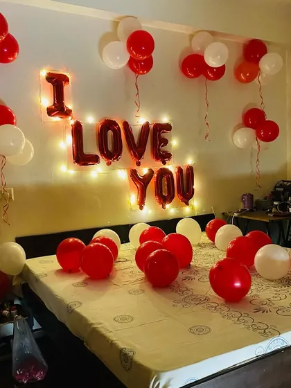 A simple and sweet "I Love You" room decoration. The setup includes red and white balloons on the bed and ceiling, with the message spelled out in foil balloons and backlit by fairy lights.