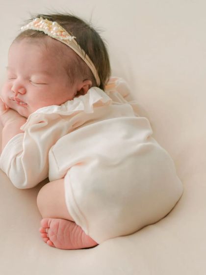 A classic newborn pose, with the baby sleeping peacefully in a simple cream outfit. This image highlights the natural, curled-up posture that is unique to the first few weeks of life.