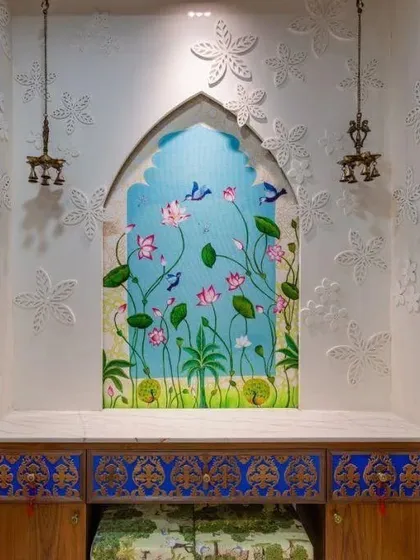 I love bringing artistic visions to life, like in this ornate niche mandir. The design features a hand-painted panel of lotuses and birds set within a traditional arch, complemented by intricate blue and gold drawers. The surrounding walls are adorned with 3D floral carvings, and classic hanging brass bells complete the temple-like feel, making it a truly bespoke piece of art.