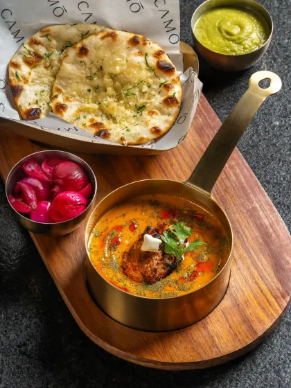A delicious chicken curry served with garlic naan for Camo Noida. The rustic wooden board and brass serving pot create a warm, inviting feel.