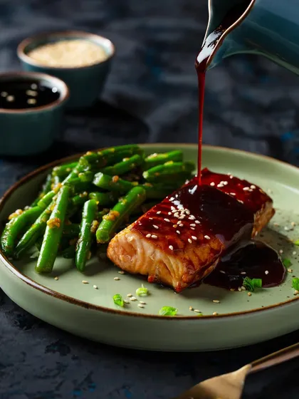 This dynamic pour shot features a rich sauce being drizzled over a piece of teriyaki salmon. The motion of the pour adds energy to the image, while the lighting accentuates the glossy texture of the sauce.