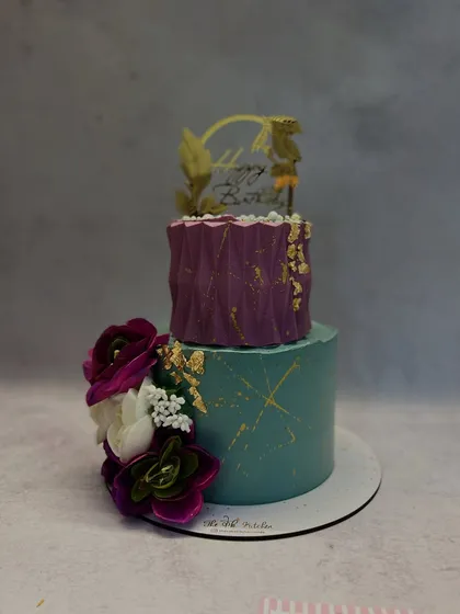 A different angle of the teal and purple cake, showing the rich colors and the way the floral arrangement cascades down the side. The 'Happy Birthday' topper makes it suitable for any stylish celebration.